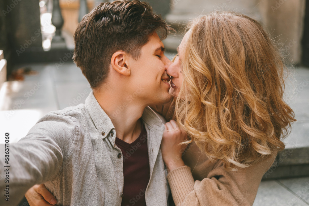 Fototapeta premium Young cheerful couple is taking a selfie while giving a kiss to each other outdoors.