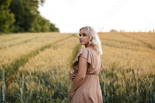 A pregnant blonde woman in a long dress is touching her belly in a wheat field at sunset. 