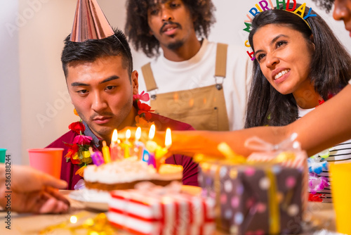 Multi-ethnic group of friends at a birthday party on the sofa at home with a cake and gifts, lighting the candles