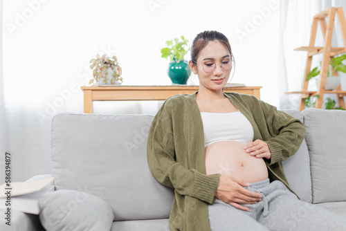 pregnant asian people sitting on sofa, she use her hand touch on her abdomen, she feeling love and care her daughter, pregnancy health care promotion