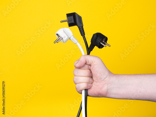 A male hand holds three electric plugs. No face, yellow background, copy space.
