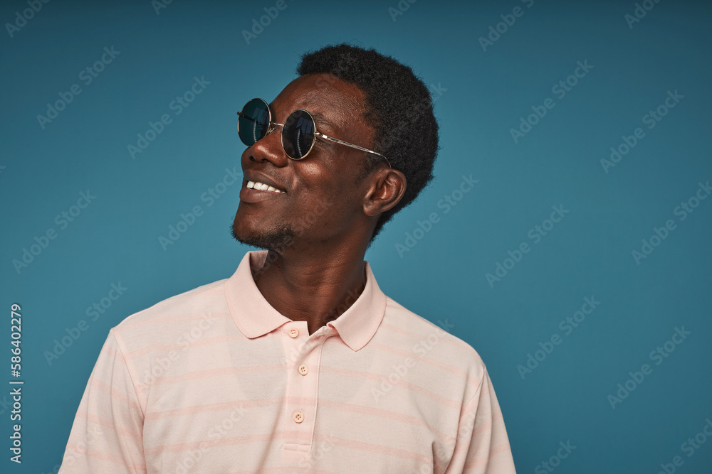Obraz premium Minimal side view portrait of smiling black man wearing sunglasses against vibrant blue background, copy space