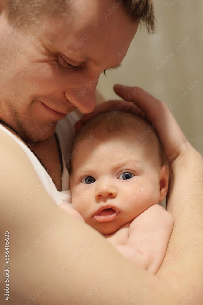 Happy father gently holds a newborn baby in his arms. The child looks into the lens. Close-up photography. Lifestyle documentary moment. Vertical photo. Happy father gently holds a newborn baby in his arms. The child looks into the lens. Close-up photography. Lifestyle documentary moment. Vertical photo.