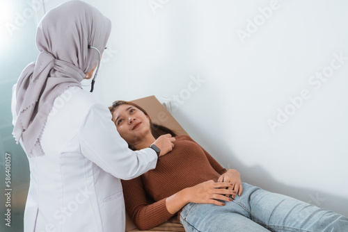 the female doctor in doctor's coat and grey veil check the female patient that laying on the bed using the stethoscope