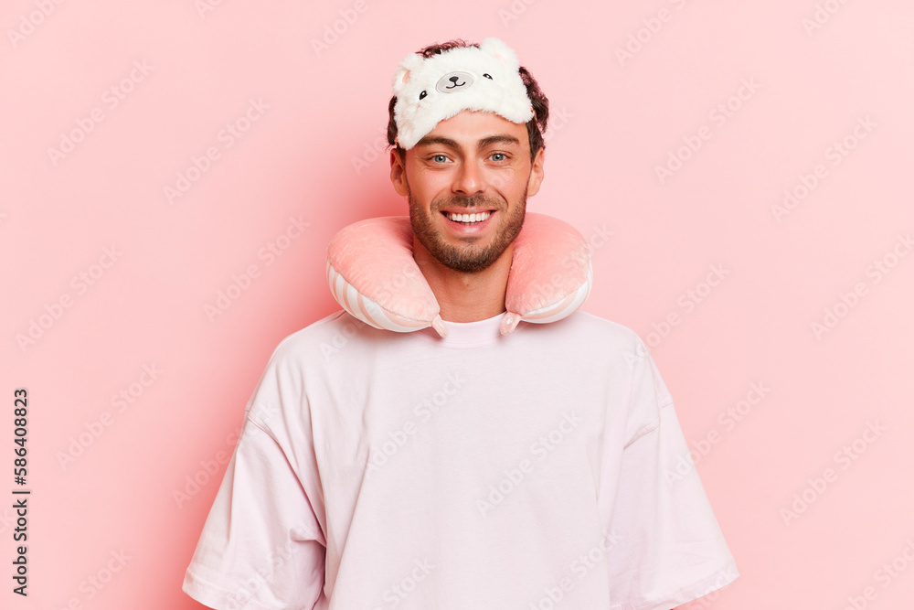 Funny bearded man in T-shirt and sleep mask on his head and sleep pillow on his neck just woke up, stands against pink background, sleep and rest concept, copy space Funny bearded man in T-shirt and sleep mask on his head and sleep pillow on his neck just woke up, stands against pink background, sleep and rest concept, copy space