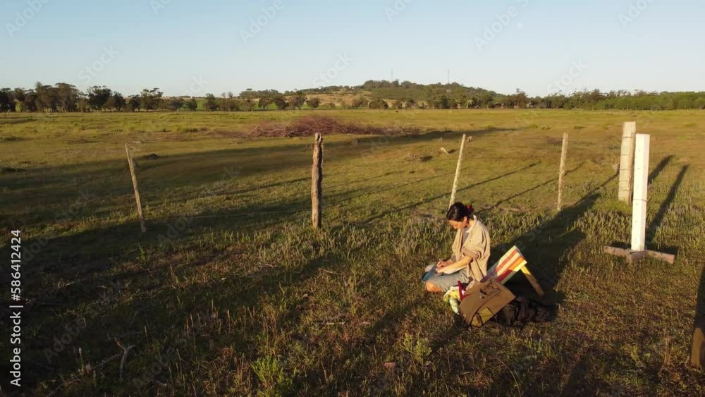 Aerial orbit shot of young woman writing and drawing on notebook outdoors in grass field during sunset - Good and free inspiration outdoors in nature Aerial orbit shot of young woman writing and drawing on notebook outdoors in grass field during sunset - Good and free inspiration outdoors in nature