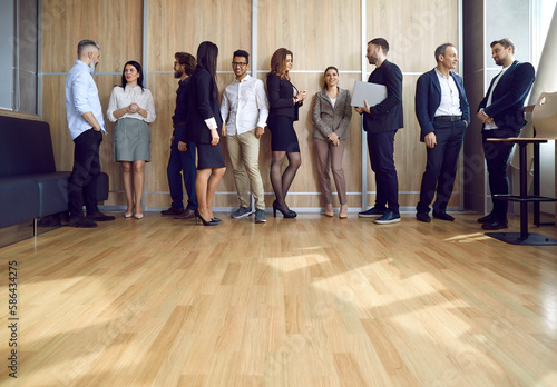 Different people communicating at corporate team meeting or business event. Multiracial men and women standing in groups in modern office with laminate flooring, talking and getting to know each other