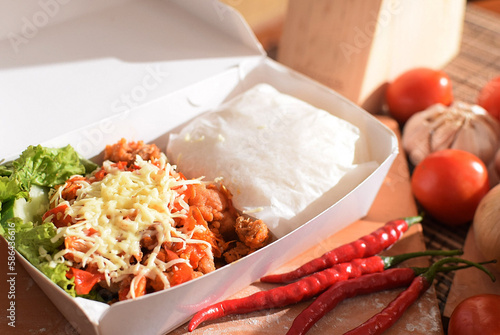 ayam geprek is indonesian spicy fried chicken with chilli sauce. served with vegetable and cheese on toping in lunch box ready to take away photo