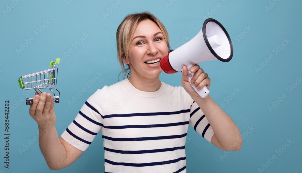 blond attractive young woman in casual attire announcing discounts using megaphone holding supermarket trolley on blue background blond attractive young woman in casual attire announcing discounts using megaphone holding supermarket trolley on blue background