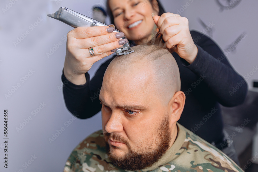 Fototapeta premium Adult man with beard in camouflage T-shirt receiving bald shave service before going to army. Girl gets rid of her hair with electric razor.
