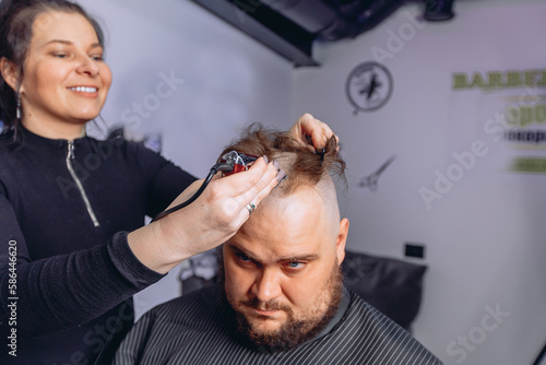 Skilled master hairdresser with smile completely shaves hair on the head of a man sitting in a chair with a cape. Provision of services in barbershop.