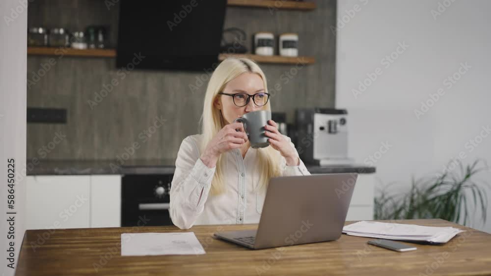 A blonde woman drinks coffee in the kitchen behind a laptop. Remote work, freelancer, work from home A blonde woman drinks coffee in the kitchen behind a laptop. Remote work, freelancer, work from home