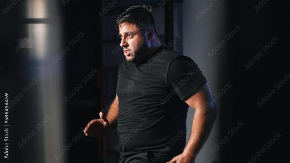 Energetic overweight man running in place as a cardio exercise for daily workout in a dark gym training hard in slow motion Energetic overweight man running in place as a cardio exercise for daily workout in a dark gym training hard in slow motion