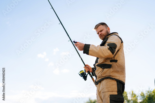 Low-angle view of fisherman holding casting rod wearing raincoat standing on bank waiting for bites on water river at summer day, looking at camera. Concept of lifestyle, leisure activity on nature.