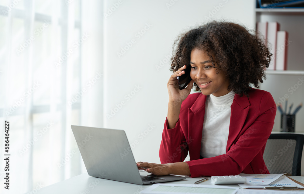 African business woman American chatting on cell phone happily chatting and reporting earnings to clients or investors working on a laptop in the office. African business woman American chatting on cell phone happily chatting and reporting earnings to clients or investors working on a laptop in the office.