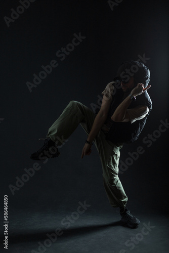 Silhouette of a male dancer dancing in the dark. Handsome fashionable man with sunglasses wearing fashionable clothes with cap, vest and sneakers dancing in motion on a black background