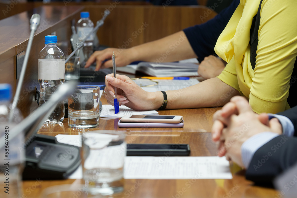 Woman, sitting at table during meeting or negotiations, holds pen and reads paper document. Hall with microphones. Lawyer, official, accountant or entrepreneur. Unrecognizable. Selective focus. Woman, sitting at table during meeting or negotiations, holds pen and reads paper document. Hall with microphones. Lawyer, official, accountant or entrepreneur. Unrecognizable. Selective focus.