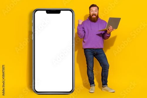 Full body photo of delighted handsome guy raise fist celebrate success hold netbook empty space panel isolated on yellow color background © deagreez