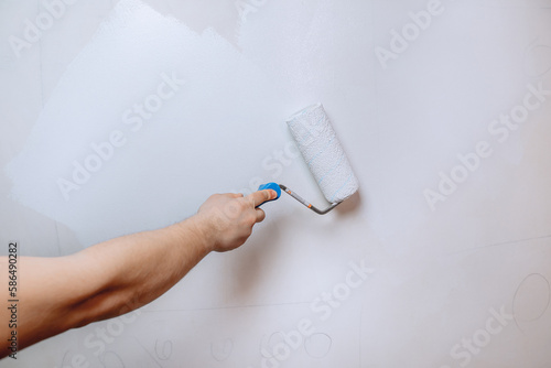 A man's hand holding a paint roller and priming the wall in white close-up. The concept of renovation in the apartment.