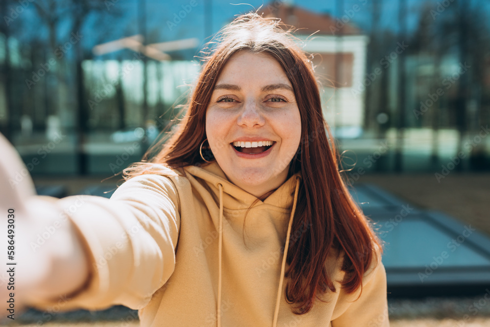 Young happy woman is making selfie on a camera on city street. Urban life concept. Girl talking on a video call with phone outdoor.