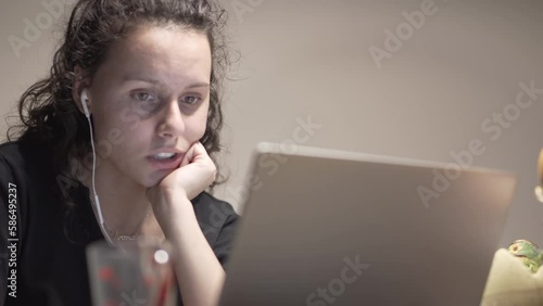 Young female IT developer wearing headphones, looking worried at laptop screen, while on an online viedeo meeting with her team mates solving an issue, working remotely at home photo