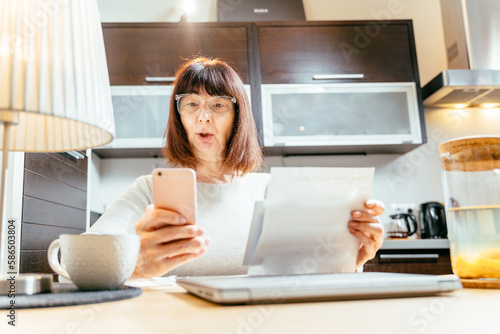 Mature Caucasian 55s woman with confused and nervous emotion sit at table calculate household finances paying bills on phone online. Excited happy woman reading message on mobile, letter in kitchen.