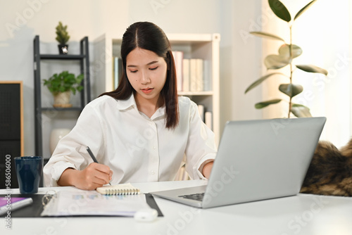 Attractive female entrepreneur sitting at desk using laptop computer and signing on business document 