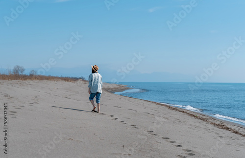 Young woman in a hat and shirt walking barefoot on a sandy sea beach leaving footprints in the sand. Back view shot