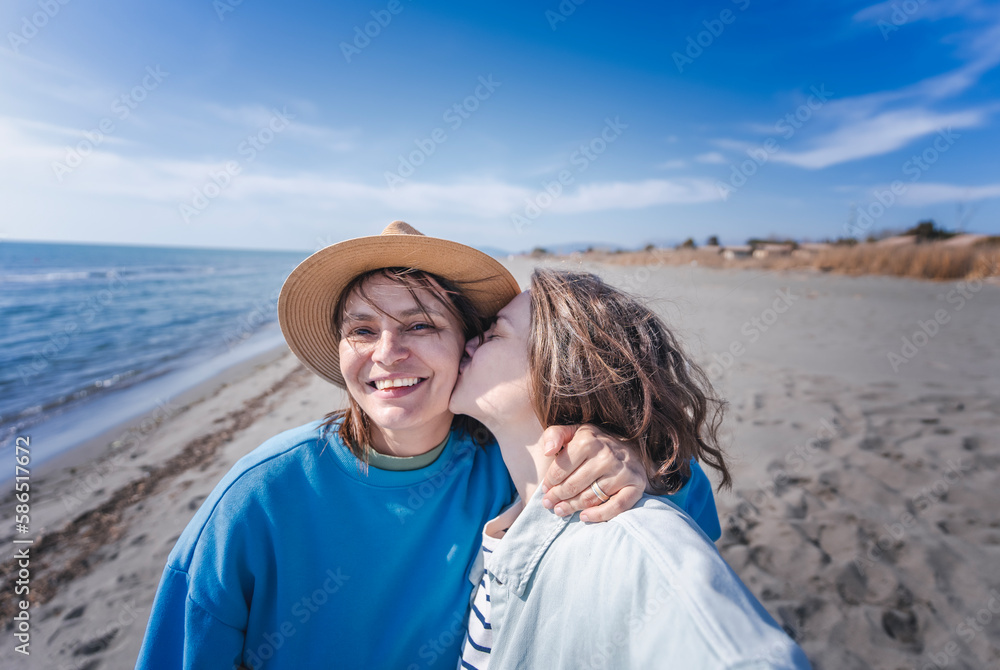 Two happy young woman lesbian couple girlfriends on sea beach during summer vacation. One kissing the other on the cheek