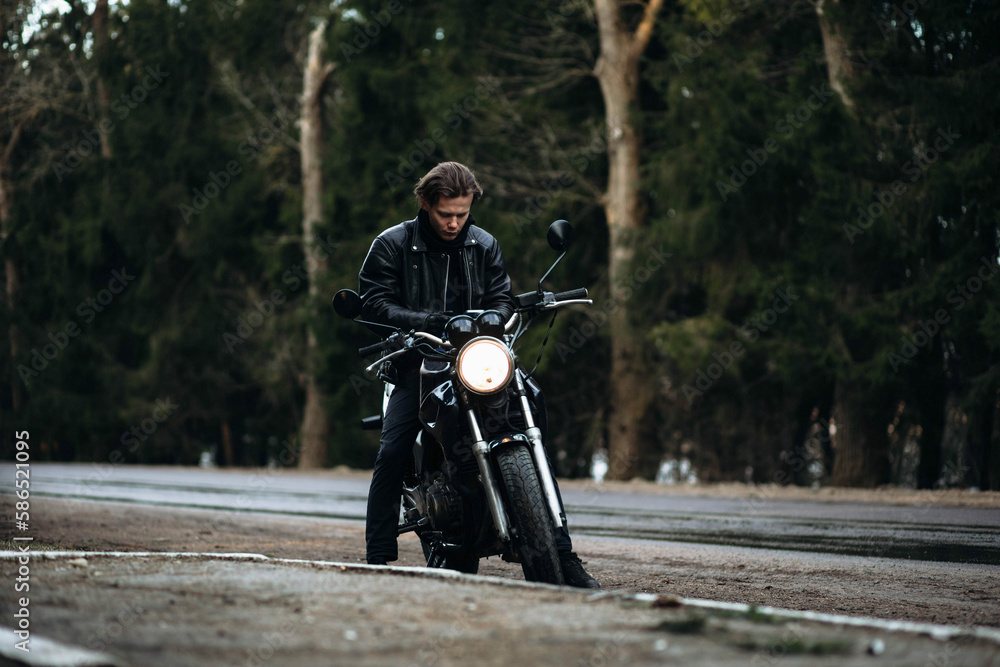 male biker motorcyclist in a leather jacket sits on a vintage motorcycle cafe racer in the forest. male biker motorcyclist in a leather jacket sits on a vintage motorcycle cafe racer in the forest.