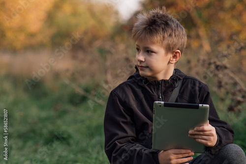 A portrait of a European blond boy in a black jacket with a tablet outside a school looking to the side for online learning, a happy young man in a park. A happy schoolboy. Autumn photo, front view