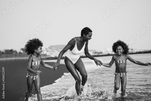 African sisters twins running on the beach with smiling mother - Focus on mom face - Black and white editing - Mom day and summer concept