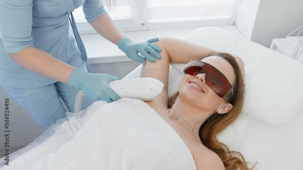 A female patient in a white cosmetic salon receives laser hair removal. Woman cosmetologist removes body hair. Professional aesthetic skin care in a beauty clinic. Smooth and soft skin hands and feet A female patient in a white cosmetic salon receives laser hair removal. Woman cosmetologist removes body hair. Professional aesthetic skin care in a beauty clinic. Smooth and soft skin hands and feet
