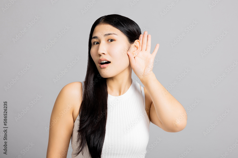Curious surprised asian girl lady woman holds hand near ear listens new interesting gossip news tries to overhear stands on grey isolated background in studio wearing basic white t-shirt. Curious surprised asian girl lady woman holds hand near ear listens new interesting gossip news tries to overhear stands on grey isolated background in studio wearing basic white t-shirt.