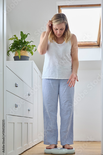 Menopausal Mature Woman Concerned With Weight Gain Standing On Scales In Bedroom At Home