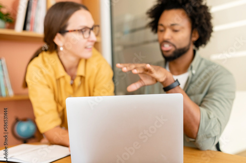 Selective focus at the laptop, two colleagues have discussion, conversation sitting side by side in modern open space on background, male mentor explaining something to colleague, brainstorming