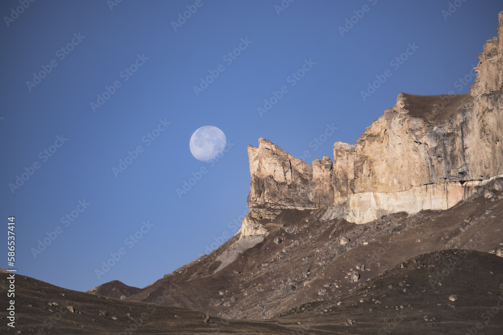 Rocky mountain peaks with the remains of snow against the background of a blue morning sky with a saturated moon, early morning in the autumn mountains