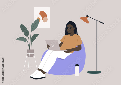 an African hipster character sits on a bean bag while typing on their laptop in their home office