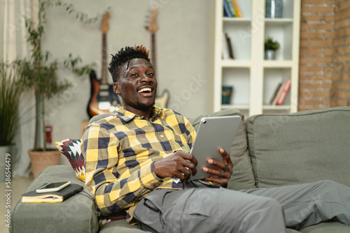 Young African man using digital tablet at home. Smiling man reading news while sitting on the couch at home.. photo