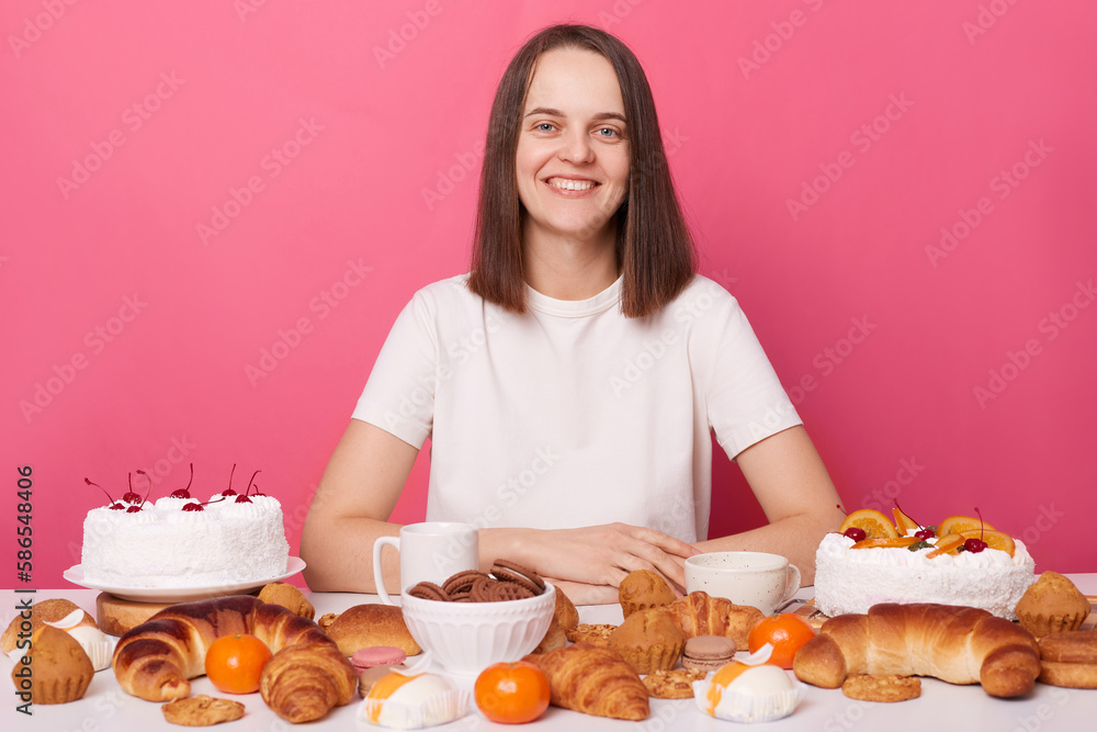 Portrait of lovely young woman with brown hair sitting at big table full of cookies, pastries and cakes, eats different desserts during holidays, posing isolated over pink background. Portrait of lovely young woman with brown hair sitting at big table full of cookies, pastries and cakes, eats different desserts during holidays, posing isolated over pink background.