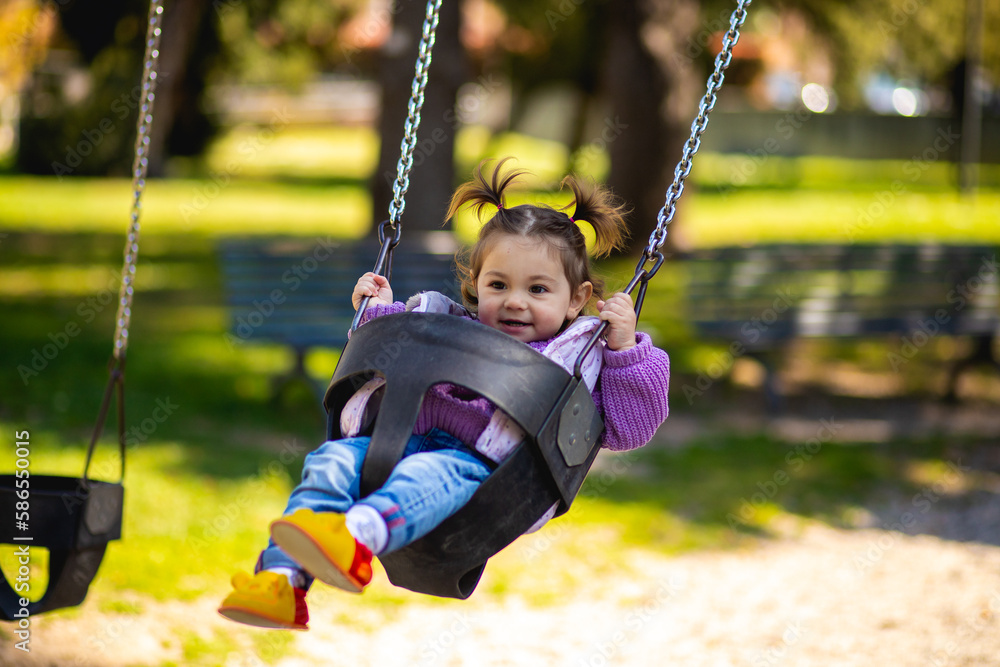 Cute caucasian little toddler girl with two tails smiling widely and swinging on a baby swing on the playground with violet sweeter sleeveless jacket and jeans