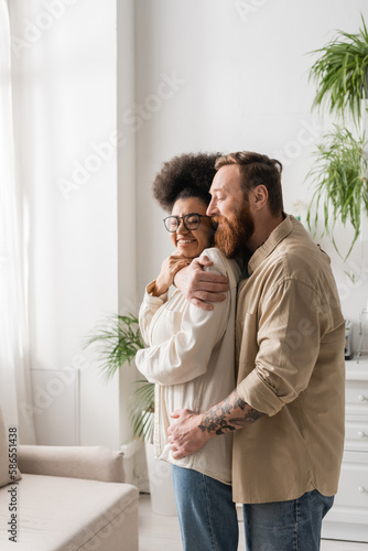 Tattooed man hugging cheerful african american girlfriend in eyeglasses at home.