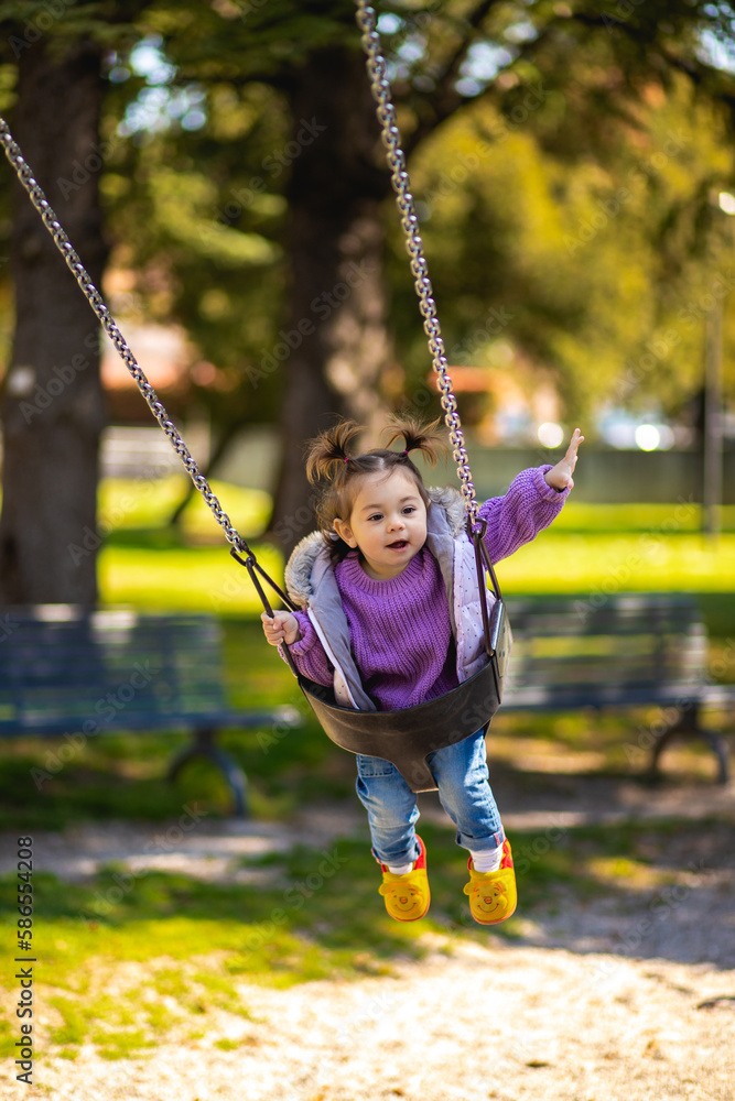 Sweet caucasian little toddler girl with two tails smiling widely swinging on a baby swing and showing he with hand on the playground with violet sweeter sleeveless jacket jeans Sweet caucasian little toddler girl with two tails smiling widely swinging on a baby swing and showing he with hand on the playground with violet sweeter sleeveless jacket jeans