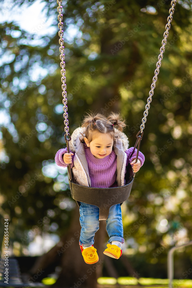 Sweet caucasian little toddler girl with two tails smiling widely and swinging on a baby swing in the playground with violet sweeter sleeveless jacket jeans Sweet caucasian little toddler girl with two tails smiling widely and swinging on a baby swing in the playground with violet sweeter sleeveless jacket jeans