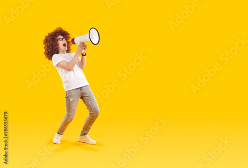 Cheerful funny preteen boy with loudspeaker in his hands makes loud advertisement on orange background. Full length child boy in curly wig announces children's sale with megaphone. Copy space. Banner. © Studio Romantic