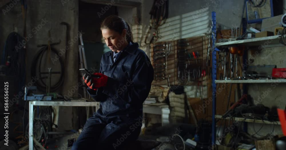Portrait of Young Female Mechanic in Uniform Overalls Using Smartphone to Chat with Family and Friends. Professional Woman Working as a Welder Taking a Break in her Repair Workshop, Staying Connected Portrait of Young Female Mechanic in Uniform Overalls Using Smartphone to Chat with Family and Friends. Professional Woman Working as a Welder Taking a Break in her Repair Workshop, Staying Connected
