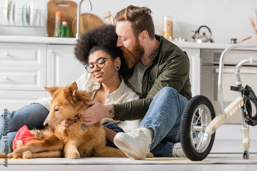 Tattooed man hugging african american girlfriend and handicapped dog near wheelchair in kitchen.
