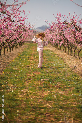 The young blondie woman in pink silk costume pink sunglasses walking and holding her straw hat into blossom peach garden 