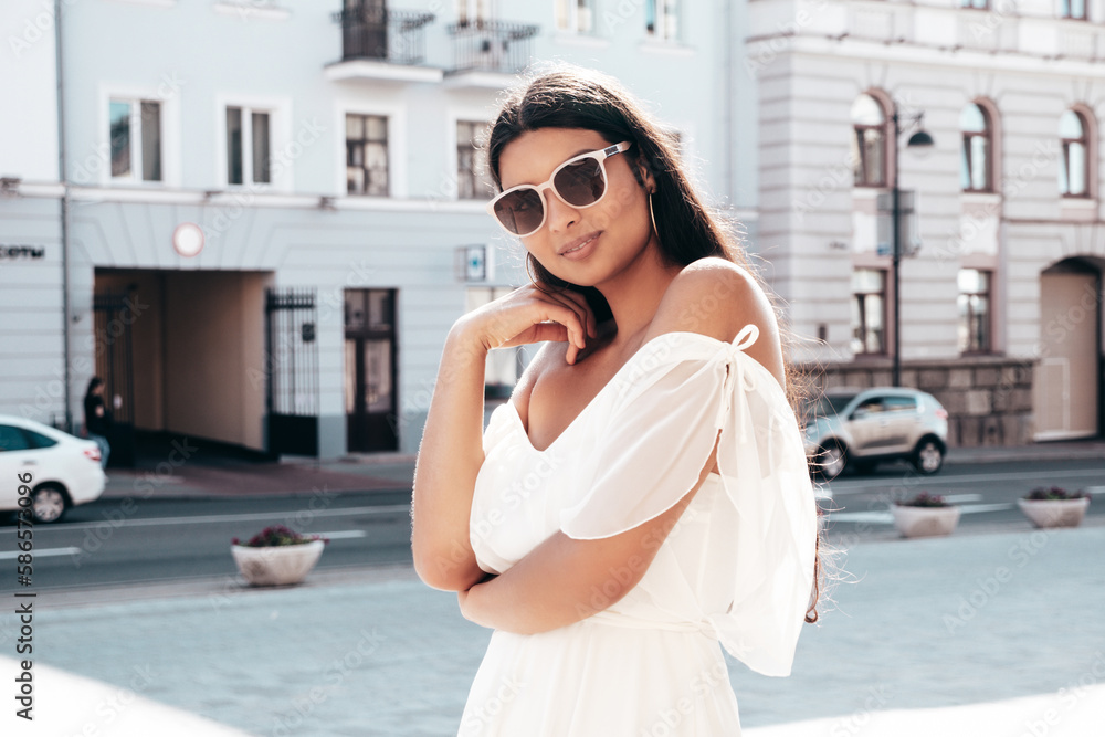 Fototapeta premium Young beautiful smiling hipster woman in white dress. Sexy carefree model posing on the street background at sunset. Positive female outdoors at warm sunny day. Cheerful and happy. In sunglasses