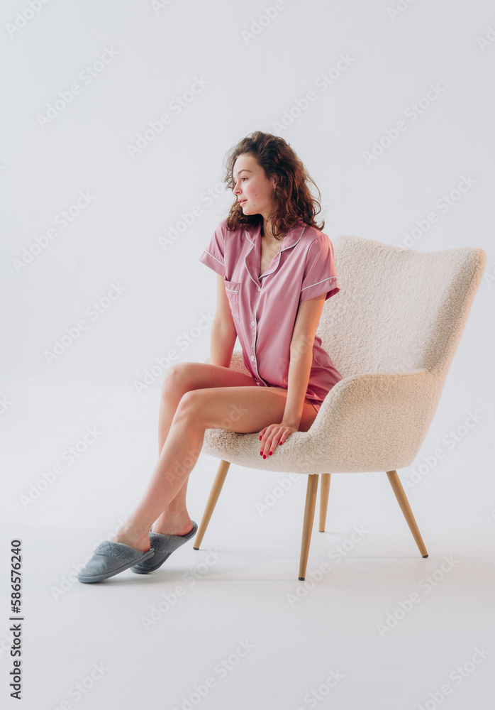 Beautiful young woman sitting in armchair on white background. Curly girl in pink silk pajama Beautiful young woman sitting in armchair on white background. Curly girl in pink silk pajama